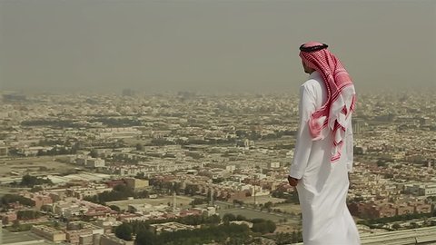 Wide shot of a Saudi Arabian man in local dress standing on a roof looking out across the city of Jeddah, In Saudi Arabia in the Middle East on a windy day.
