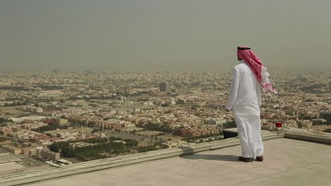 Wide shot of a Saudi Arabian man in local dress standing on a roof looking out across the city of Jeddah, In Saudi Arabia in the Middle East on a windy day.
