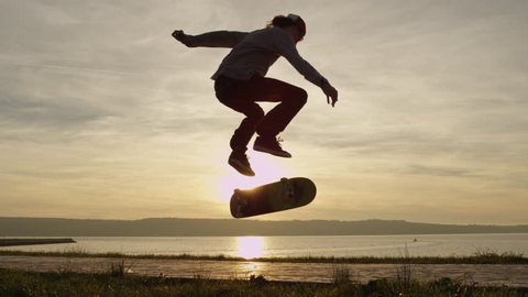 SLOW MOTION CLOSE UP, DOF: Silhouetted skateboarder skateboarding and jumping kickflip trick on walking path along the ocean coastline against golden setting sun. Skater riding skateboard doing tricks