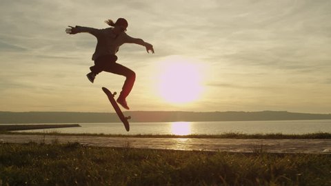 SLOW MOTION, CLOSE UP: Silhouetted skateboarder skateboarding and jumping hardflip trick on boulevard along the ocean at golden light sunset. Skateboarder riding skateboard at sunrise at seaside