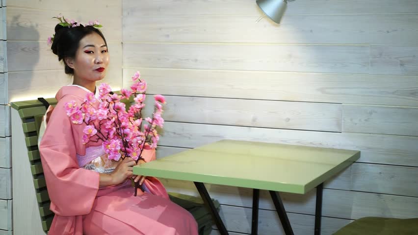 Beautiful geisha in traditional Japanese kimono pink sitting at an empty table in the restaurant and picks at the branches of cherry blossoms.