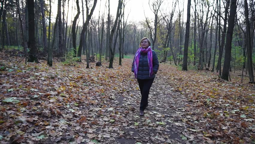 Senior woman with short hair, glasses on her eyes, the warmth dressed walking in the autumn woods, where the trees do not have leaves, it fell from the branches, and is on track in the park.