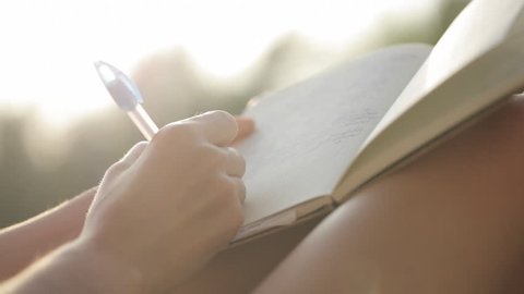 Young woman sitting at balcony in sunlight and writing in diary with garden of palm trees on the background