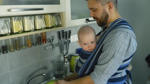 Young father with his son in sling washing dishes
