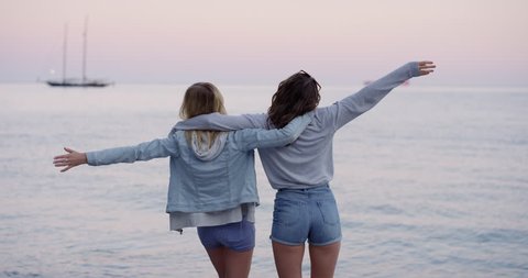 Best friends lifting arms up on beach watching sunset over ocean looking at pink sky wearing denim summer shorts on road trip adventure