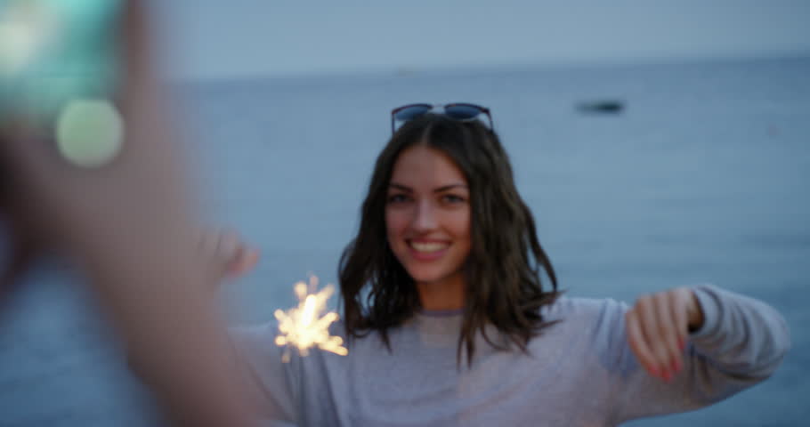 Young woman taking photo of best friend holding sparklers dancing in slow motion celebrating new years eve and independence day with fireworks at sunset on beach with ocean background