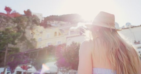 Beautiful tourist woman looking up at view of Positano Amalfi Coast low angle shot with warm sun flare enjoying Italy vacation travel adventure wearing cute dress and straw hat