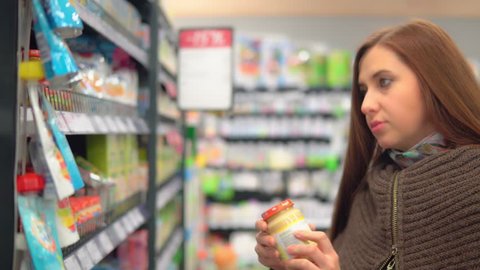 A young attractive woman shopping in a supermarket, taking baby food banks off the shelf, viewing, reading the inscriptions on the labels,comparing and choosing. 4K
