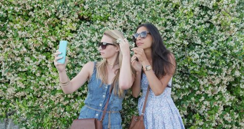 Best friends on holiday taking selfie using smartphone wearing flower in hair. Girls sharing lifestyle photo using mobile phone standing in front of wall of flowers sending message enjoying summer
