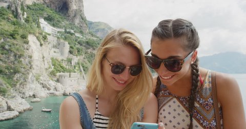 Two young women hanging out at beach using mobile phone sending text message snapchat sharing digital content on social media tourist girls enjoying summer vacation on Amalfi Coast Italy