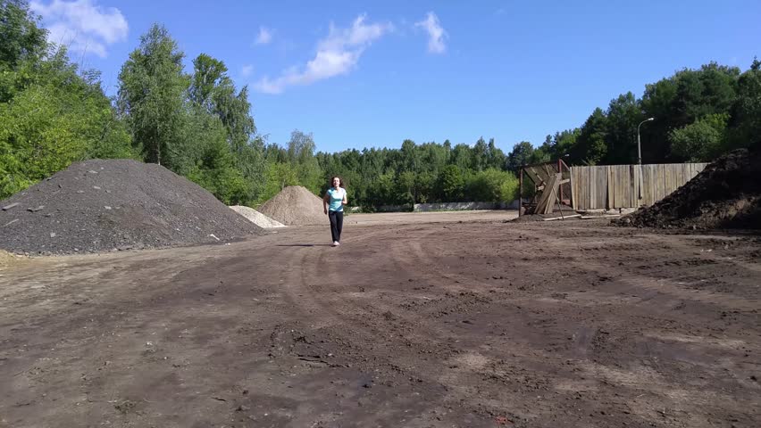 Woman in blue shirt run on ground near hills of asphalt bits and park at sunny day, mobile phone video.