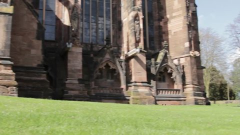Panning Shot showing Lichfield cathedral in all its Gothic Grandeur from foundations to spire, weatherworn stone, standing firm against a blue sky and lush green grass in the small city of Lichfield.
