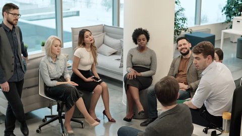 High angle shot of group of businesspeople sitting in circle in modern office and listening to male colleague telling story during meeting or team building exercise