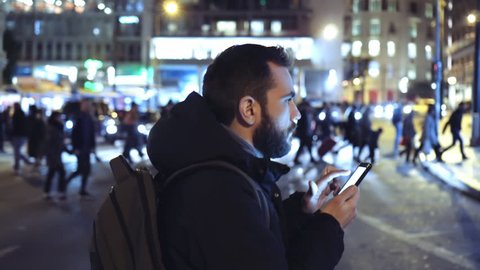A young man is commuting to work while browsing his cellphone on a busy urban street at night.