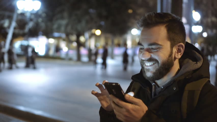 A smiling cheerful young man is commuting to work while browsing his cellphone on a busy urban street at night. 100fps slowmotion. Royalty-Free Stock Footage #24940649