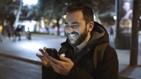 A smiling cheerful young man is commuting to work while browsing his cellphone on a busy urban street at night. 100fps slowmotion.