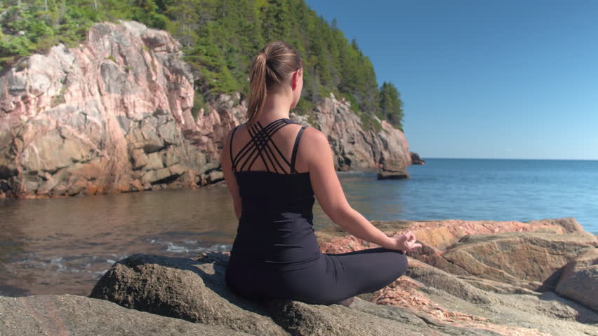 CLOSE UP: Attractive young woman meditating sitting on red volcanic rocky shore and enjoying relaxing sunny spring day. Girl relaxing doing meditation by the seashore in lush overgrown wilderness