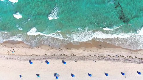 Video loop with aerial footage showing bird eye view with people walking and getting tan on a tropical Florida beach under blue umbrellas and turquoise ocean waves breaking against the coastal line