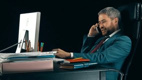 Cheerful bearded businessman working on his computer and talking on cell phone. Black background. 4K video