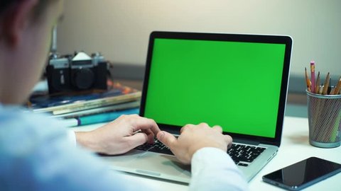 A man types on a laptop on his desk. Green screen for your custom screen content.
