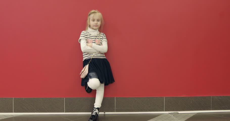 4K A stylish little blond girl leaned against the red wall. A fashionable beautiful girl in a dress is standing in the hall.