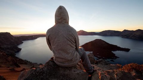 2.5D parallax video hooded man sitting on edge of rock cliff watching sunrise above crater lake, Oregon, USA