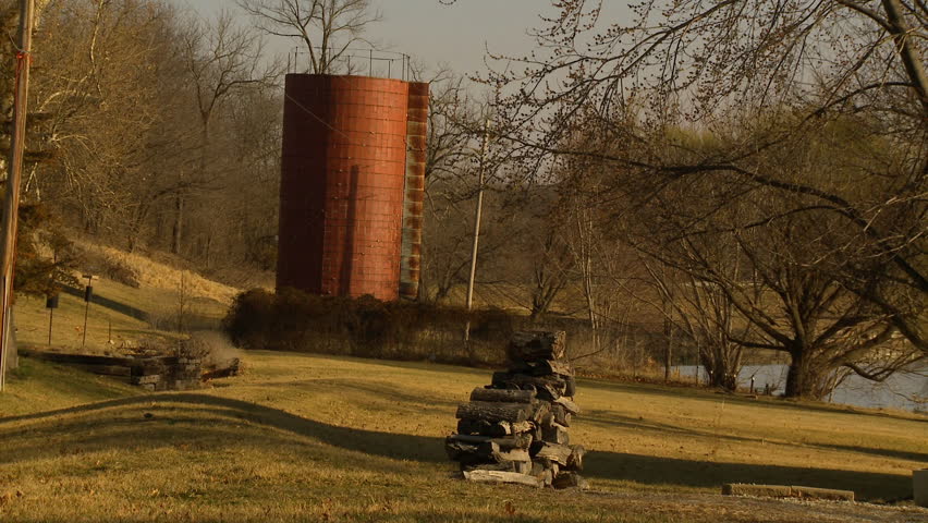 An old farm grain silo stands near a lake with trees and a fire wood pile on a spring day in Kansas. Rural America.