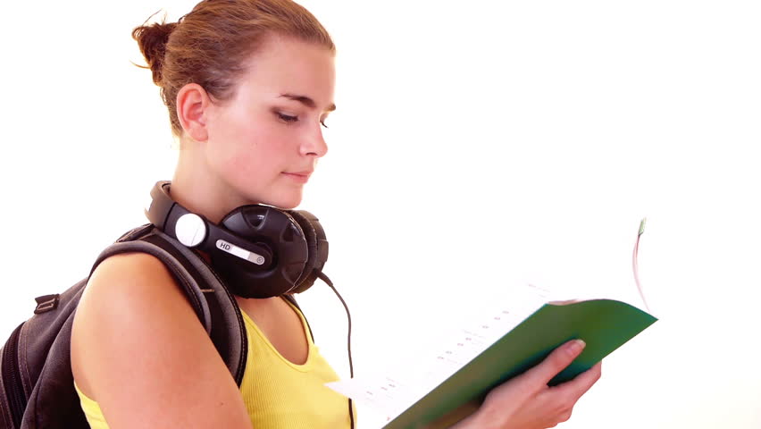 Smiling schoolgirl with headphones and backpack reading something in textbook 