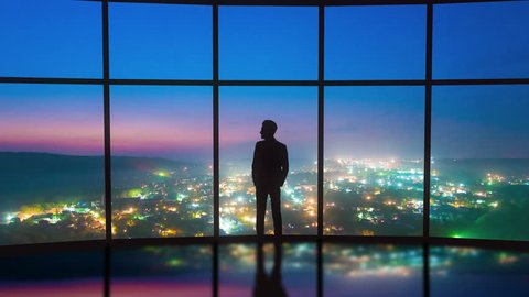 The man stand near the panoramic window on the night city background. Time lapse