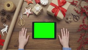Aerial lockdown footage of woman using digital tablet on table. Personal perspective of female touching green screen on device. Overhead flat lay of gift wrapping equipment on table during Christmas.