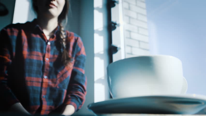 Beautiful girl hippy takes a mug of coffee from a vintage wooden blue table. She is dressed in a plaid red shirt, a hipster style. A large window, the sun shines brightly