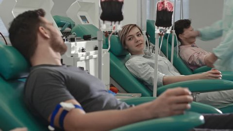 Tracking shot of young woman and man sitting in medical chairs and chatting while donating blood in hospital