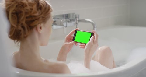 Woman using smartphone touchscreen chroma-key greenscreen enjoying relaxing bubble bath close-up sharing authentic social media on digital display