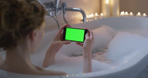 Woman using smartphone touchscreen chroma-key greenscreen enjoying relaxing bubble bath close-up sharing authentic social media on digital display