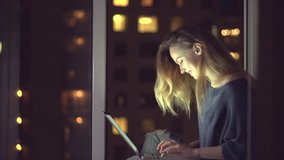 Beautiful Girl sitting near the window at home at night and using laptop. Nighttime working. Chatting young woman in dark room. 4K UHD video