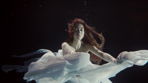 young woman with long red hair swimming underwater in a white dress like a fairy tale