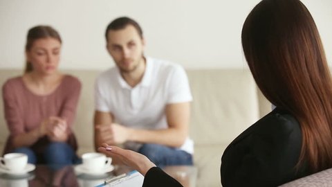 Young upset couple visiting psychotherapist office, listening to marriage counselor advice, looking at each other, making up after therapy session. Focus on therapist back who is talking and gesturing