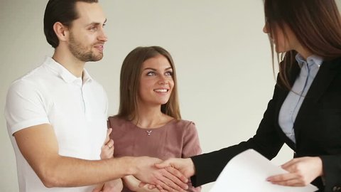 Young attractive happy smiling couple meeting real-estate agent. Overjoyed husband and wife choosing and buying new house, shaking hands with female broker, getting keys and embracing each other