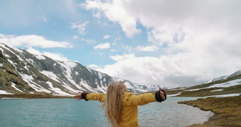 Woman with arms raised by lake looking at snowy mountain view lifting arm up celebrating scenic landscape Girl wearing yellow jacket enjoying vacation travel adventure nature Norway
