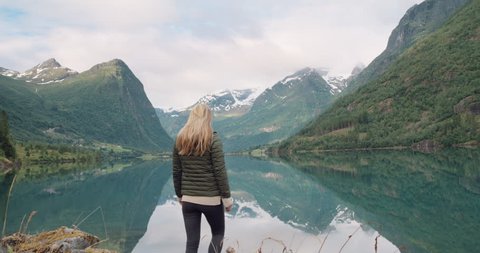 Woman with arms raised standing at edge of lake looking mountain view reflection Hike girl wearing green down jacket lifting arm up celebrating landscape enjoying vacation travel adventure Norway