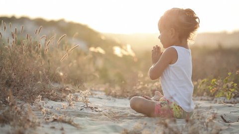 Little cute girl in white shirt meditates at sunset sitting on the sand light warm wind