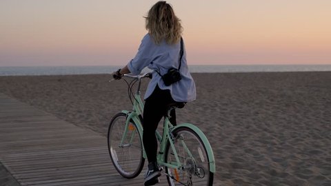 Pretty cute blonde female rides her mint hipster bike on the beach with her hair flowing in the wind and makes a photo of the sunset while dreaming in her thoughts.