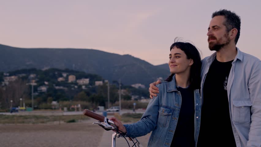 Cute trendy hipster couple in casual clothes is cuddling and hugging on the beach watching pink sunset, girl holds a mint bike
