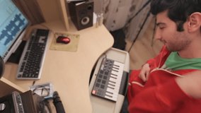 Young man sitting in front of computer and midi keyboard. He listens to music and waving his arms. Top view.