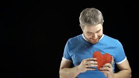 Happy man in blue tshirt holds one red heart shape. Love, single, romance, dating, relationship concepts. Black background. 4K video
