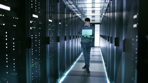 Male Server Engineer Walks with Laptop Through Working Data Center Full of Rack Servers. Shot on RED EPIC-W 8K Helium Cinema Camera.