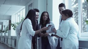 Four diverse students in white coats standing in medical university hall near big window, holding books, and having a discussion and using smartphones