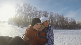 Funny sister and brother are sledding in the mountain through the sun in slow motion on sunny winter day and recording themself on video with action camera. 1920x1080