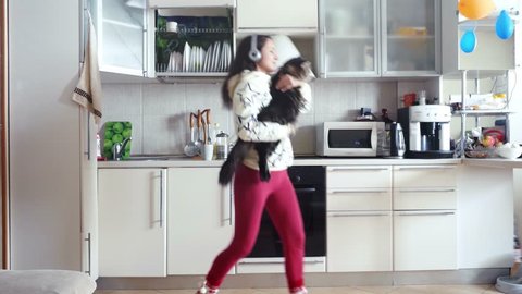 Joyful young beautiful woman is dancing in kitchen with her adorable Maine Coon cat wearing headphones in the morning listening to music on smartphone. 3840x2160
