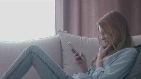 Attractive blonde Caucasian teen girl sitting on a sofa at home and texting on her phone. Smiling, laughing female. Love, friendship concept. 4K UHD RAW edited footage